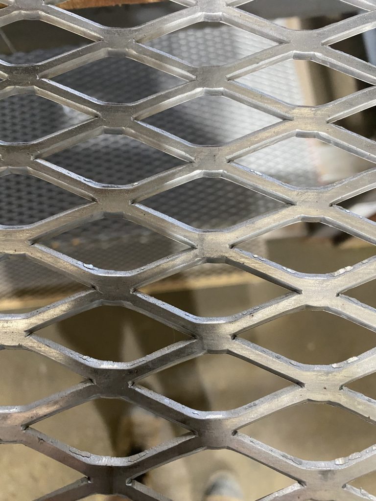 This is flattened, non-galvanized expanded metal grating. The sharp edges inside the diamond shape openings indicate slitting. The diamond shape openings and thick strands make cuts difficult for a novice equipped with poor quality grinder and substandard cutting disk due to excessive vibration when jumping from one diamond to the next. By JW Investrade Inc. for metalgrating.ca, 2022, London, Ontario