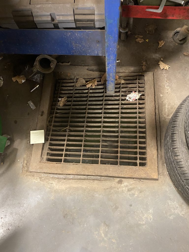 Old floor drain cover made from reversed steel angle rim and steel metal grating has collapsed in the upper left corner under heavy load of steel shelf's leg. The welds between the steel grating and the rim broke off. JW Investrade Inc. for metalgrating.ca 2023, London, Ontario.