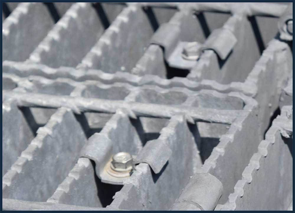 Saddle clips on grate steel for pedestrians. The saddle clip bridges the 2 bearing bars and then fasten them to supporting grate steel panel's flange. The saddle clip uses number of different fasteners that allow the attachment of the clip to supporting flange. JW Investrade Inc for metalgrating.ca 2024 London Ontario