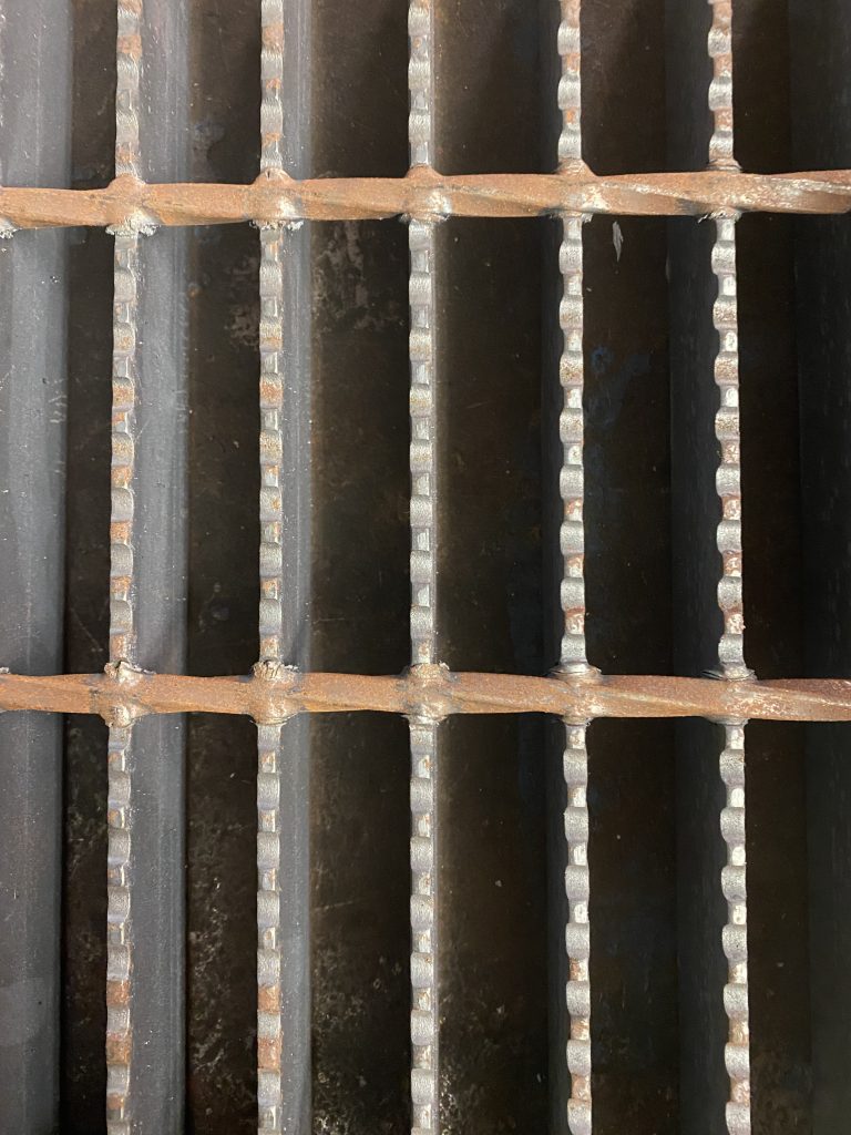 Closeup structure of Closeup of unpainted and non galvanized, serrated welded bar grating with clearly visible welds. The cross bars consist of twisted round bars which are positioned on top of load bearing serrated flat bars. By JW Investrade Inc. for metalgrating.ca 2023, London, Ontario.