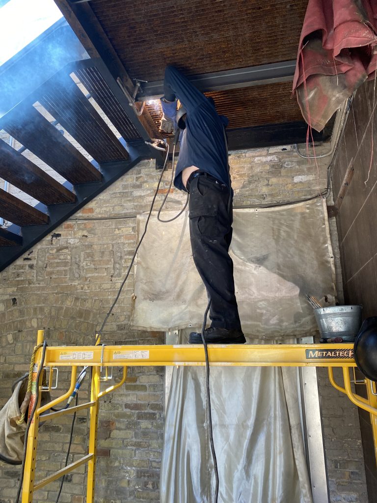 Welder weds in the first supporting structural steel channel to heavy beam frame under the grate steel deck. The first supporting channel is placed under the small gap between first and second grating panel. JW Investrade Inc. for metalgrating.ca 2021, London, Ontario.