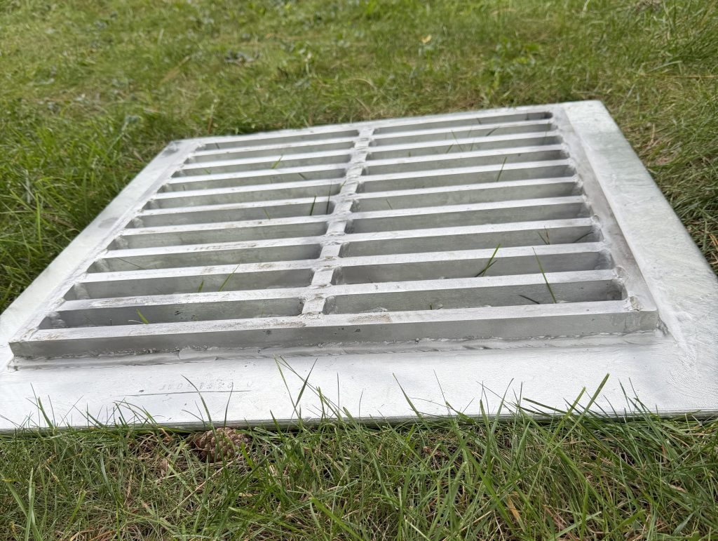 The upside down and angular view of unsupported style lid exposes steel plate's support, custom steel grating, welded to the steel plate. the height of the support is 1". The flanges of steel plate not cover by steel grating allow for hanging effect of this lid. For JW Investrade Inc.www.metalgrating.ca, London, Ontario. 2026