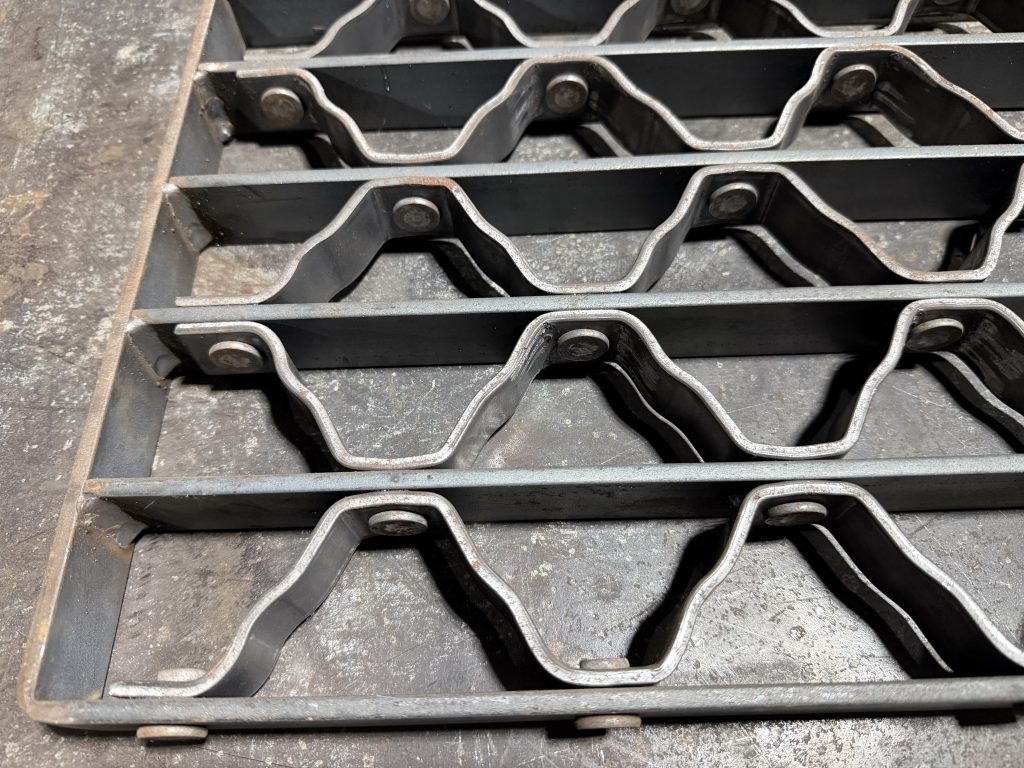 Riveted grating is a heavy-duty, structurally robust form of industrial metal bar grating made by joining straight bearing bars and bent or crimped connecting reticulated bars with cold-pressed rivets. It is perfect for bridges and overpasses. Rivetted grating is very expensive grating that is not very useful for trenches and driveways due to its complex structure and high cost. JW Investrade Inc. for Metalgrating.ca, 2026, Lucan Ontario